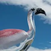 Glass flamingo sculpture, clear with pink mottling, long curved neck and legs, set against a blue sky and white clouds.