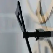 Stainless steel arbor, “takto presto,” with cylindrical support, black angular arms, and cut-out notches, against blurred mechanical gears.
