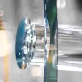 Chrome-edged circular glass component with blue-green iridescence, paired with a vertical chrome element, against a blurred reflective backdrop.