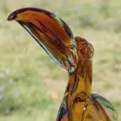 Glass toucan sculpture, amber with brown swirls, featuring a curved beak and transparent details against a blurred floral background.
