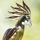 Polished metal parrot sculpture with green-to-gold gradient, fanned wings, ruby accents, and detailed beak against a blurred green backdrop.