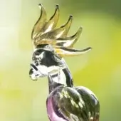 Glass parrot sculpture, transparent with purple & green tints, featuring a golden, curved crest and detailed beak, set against a blurred green backdrop.