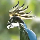 Glass parrot sculpture, turquoise and blue, with black head, white eye patch, and golden-yellow crest plumes against blurred green.