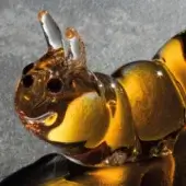 Glass caterpillar sculpture, amber-colored with swirling patterns, curled form, glossy finish, dark eyes, and textured gray surface.