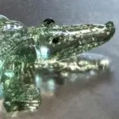 Green glass alligator sculpture, translucent texture, realistic form, detailed teeth, reflective dark gray surface, decorative art object.