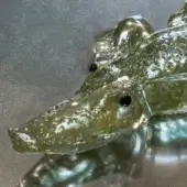Glass crocodile sculpture, translucent green-gold, textured skin detail, elongated form with pointed snout and dark circular eyes.