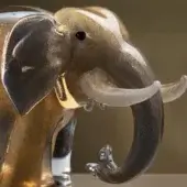 Glass elephant sculpture, mottled grey and gold, stylized form with curved trunk and single ivory tusk on clear glass base.