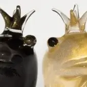 Glass frog sculptures, black with gold flecks and translucent gold, feature elongated bodies and spiked crowns against a white backdrop.