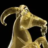 Glass sculpture, gold-colored, depicting a rearing goat with curved horns and textured, speckled finish against a black backdrop.