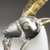 Glass goat head sculpture, clear with gold leaf horns, featuring swirling patterns and a dark gray muzzle against a gray backdrop.