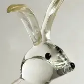 Glass rabbit sculpture with white body, black eyes, and amber-tipped ears holds a clear glass carrot against a gray backdrop.