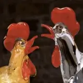 Glass rooster sculptures, gold and transparent with red accents, stylized design, elongated necks, displayed against a dark brick wall.