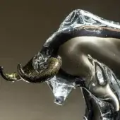 Glass ox sculpture, clear and black, with gold speckles on curved horns, dynamic form against a brown backdrop.