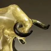 Blown glass ox sculpture, translucent amber with golden shimmer, featuring textured black and gold speckled horns, simplified abstract form.