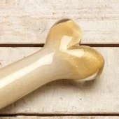 Ceramic bone sculpture, creamy white to gold glaze, stylized femur shape with glossy finish on weathered wood planks.