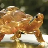 Glass tortoise sculpture, amber and gold, detailed shell and legs in walking pose, translucent with black eyes, blurred foliage background.