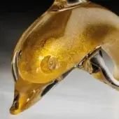 Golden glass dolphin sculpture, translucent form with gold flecks, stylized body, clear dorsal fin, reflective white surface.