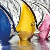 Glass fish sculptures, blue, amber, and ruby, with elongated bodies and curved fins, displayed on a reflective surface against silver foil.