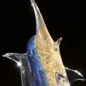Glass marlin sculpture, blue and gold layered design, translucent form with defined snout and crystalline fins against black backdrop.