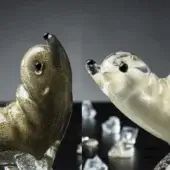Glass seal sculptures on clear ball bases, mottled golden-brown and clear glass, black bead eyes, dark grey background, studio lighting.