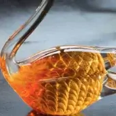 Glass swan sculpture, amber and gold tones, textured scale-like pattern, iridescent finish, studio lighting, dark surface display.