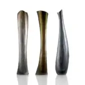 Glass vases, smoky gray, golden amber, charcoal – elongated forms with vertical striations and sculpted silhouettes against a white backdrop.