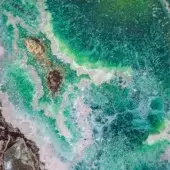 Turbulent ocean water crashes against dark rocks, displaying swirling teal, emerald, and blue currents with white foam and light reflections.