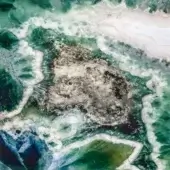 Sicis Vetrite surface, green boulder texture, irregular rounded shapes, turquoise water, sandy shoreline, layered sedimentary material.