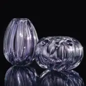 Glass vessels, polished amethyst, feature ribbed egg shape and rounded organic form, reflecting light against a black backdrop.