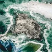 Dark gray boulder amidst turbulent turquoise and teal water, featuring a rough, circular-patterned surface and white foam waves.