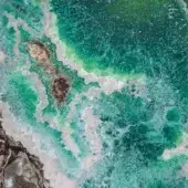 Turquoise ocean water crashes against dark, jagged rocks with white foam, displaying swirling currents and wave formations in a cool, vibrant scene.