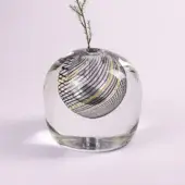 Glass vase, clear with black and gold filigree sphere inside, rounded form, lavender backdrop, and small evergreen sprig.