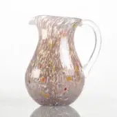 Glass pitcher with colorful flecks, rounded body, clear handle, and angled spout on a white reflective surface.
