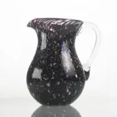 Black glass pitcher with clear handle, speckled with red, yellow, green, blue, and white fragments, rounded body, flared spout, glossy finish.