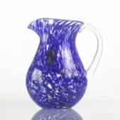Blue glass pitcher with clear handle, rounded body, and speckled iridescent glass inclusions, centered on a white reflective surface.