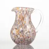 Glass pitcher with multicolored speckled pattern, rounded form, flared spout, and curved handle on a white reflective surface.