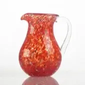 Red glass pitcher with speckled pattern, rounded body, and flared spout reflecting on a white glossy surface.