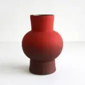 Red ceramic vase with rounded, bulbous shape and matte finish displayed on a white surface against a neutral backdrop.