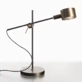Desk lamp, anodic bronze, cylindrical shade with slender stem on gray surface, minimalist geometric design with black cord and circular knob.