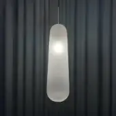 Suspension lamp, frosted glass, elongated teardrop shape with ribbed texture, silver metal rod, centered against dark gray pleated curtains.