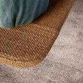 Textile bed frame, golden-brown woven material, repeating nubby pattern, with teal velvet pillow and adjacent gray rug, studio lighting.