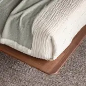 Wood bed frame, medium-brown finish, rounded edge, with sage green fringed blanket and creamy linen-textured throw, on neutral carpet.