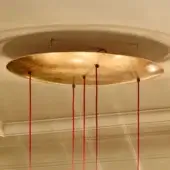 Circular gold canopy with red suspension cords hangs from a beige ceiling with recessed molding and panelled walls in softly lit room.