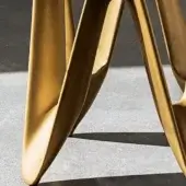 Sculptural table base, burnished gold metal, featuring flowing curves and three intersecting support arms on a concrete floor.