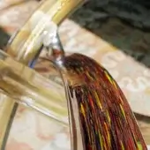 Multicolor glass vessel with red, yellow, black, and blue murrine streaks held by a curved gold metal stand, background includes blurred currency.