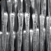 Transparent crystalline forms, vertically suspended, display ridges and grooves with intense light reflection in grayscale tones.