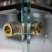 Gold faucet handles with cylindrical bodies are reflected in clear glass panes, framed by dark metal, showcasing a polished gold finish.