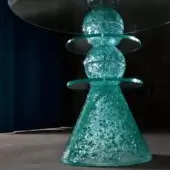 Glass table, turquoise textured base, multi-tiered conical form; clear glass top, stacked fractured glass elements, teal curtain backdrop.
