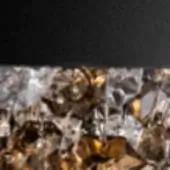 Caramel candies in silver foil, golden-brown and amber tones, densely packed against a dark gray background, creating a textured, reflective surface.