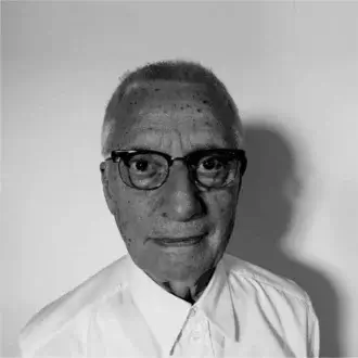 Elderly person with sparse hair and glasses looks forward with a slight smile wearing a light collared shirt against a plain background